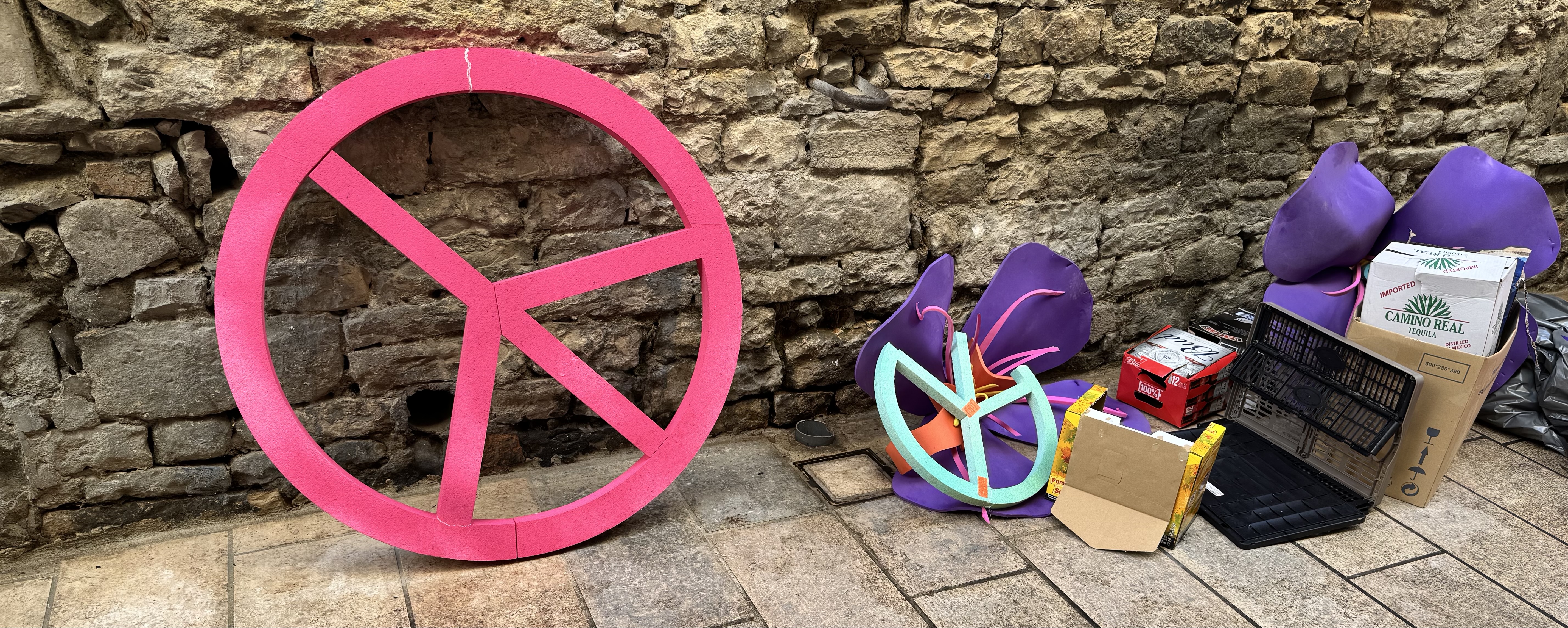 Large pink peace symbol leaning against a stone wall, alongside a collection of discarded items including purple foam shapes, a smaller multicolored peace symbol, cardboard boxes (one labeled "CAMINO REAL TEQUILA"), plastic crates, and packaging materials arranged on tiled pavement.