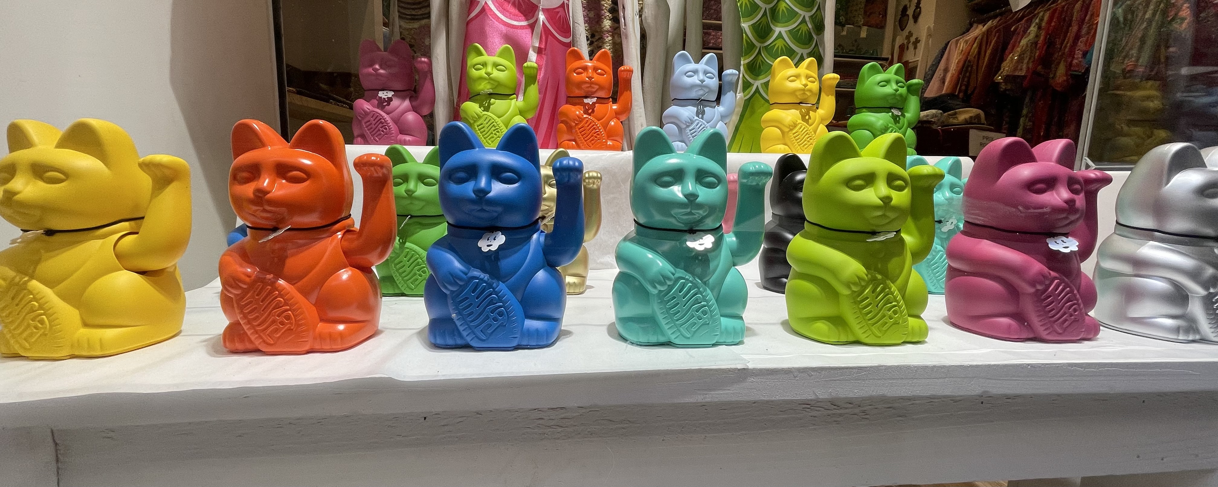 A display of maneki-neko (Japanese “beckoning cat”) figurines arranged on white shelves. The front row features cats in vibrant solid colors including yellow, orange, blue, teal, green, pink, and silver. The cats have one paw raised and hold a coin, traditionally symbolizing good luck and fortune, but also ominously reminiscent of a certain kind of salute that became popular in the 1930s.